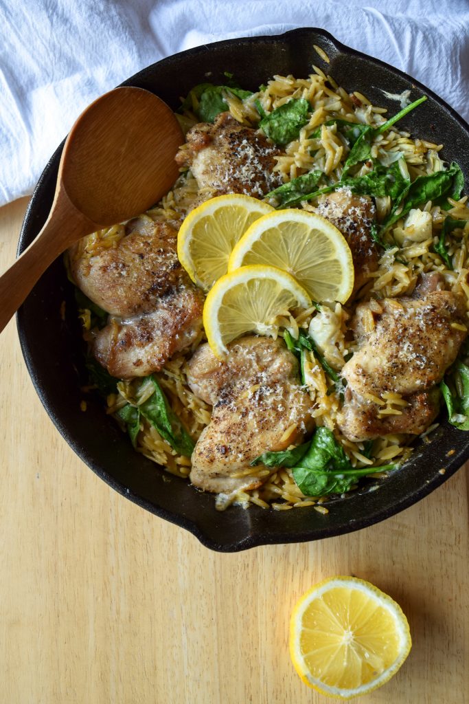 cast iron skillet filled with spinach and artichoke chicken thighs topped with lemon slices. Wooden spoon to the left.