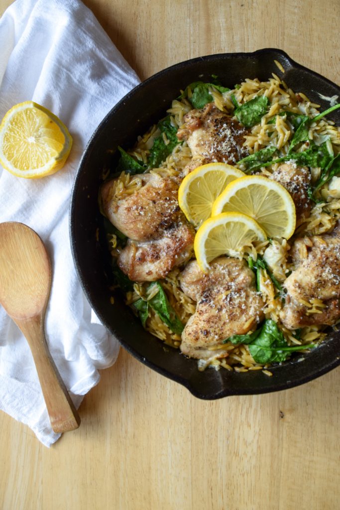 cheesy spinach and artichoke chicken thighs in orzo in a cast iron skillet, topped with 3 sliced lemons. To the left, a white linen towel with a wooden spoon and a lemon.