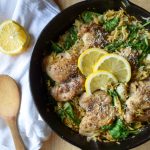 cheesy spinach and artichoke chicken thighs in orzo in a cast iron skillet, topped with 3 sliced lemons. To the left, a white linen towel with a wooden spoon and a lemon.