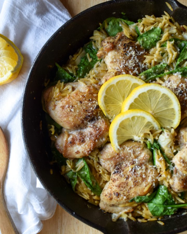 cheesy spinach and artichoke chicken thighs in orzo in a cast iron skillet, topped with 3 sliced lemons. To the left, a white linen towel with a wooden spoon and a lemon.