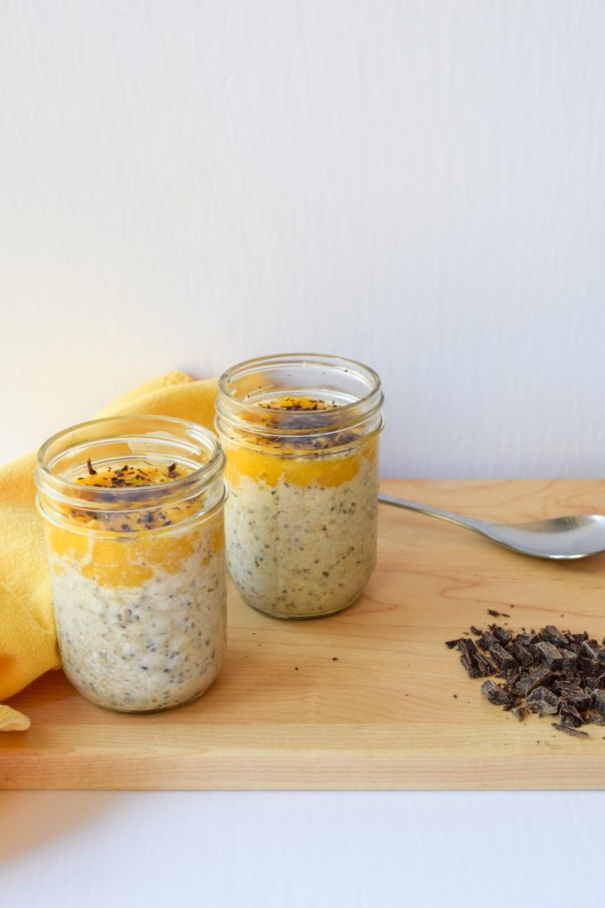 2 small mason jars filled with overnight oats, topped with mango coulis and dark chocolate atop a wooden cutting board.