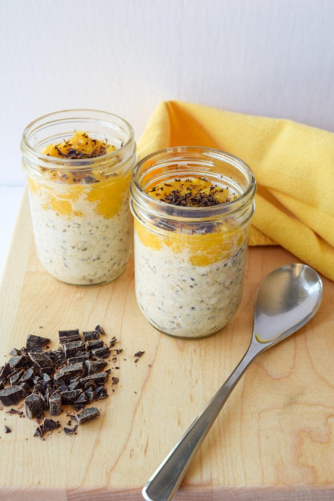 2 small mason jars filled with overnight oats, topped with mango coulis and dark chocolate atop a wooden cutting board.