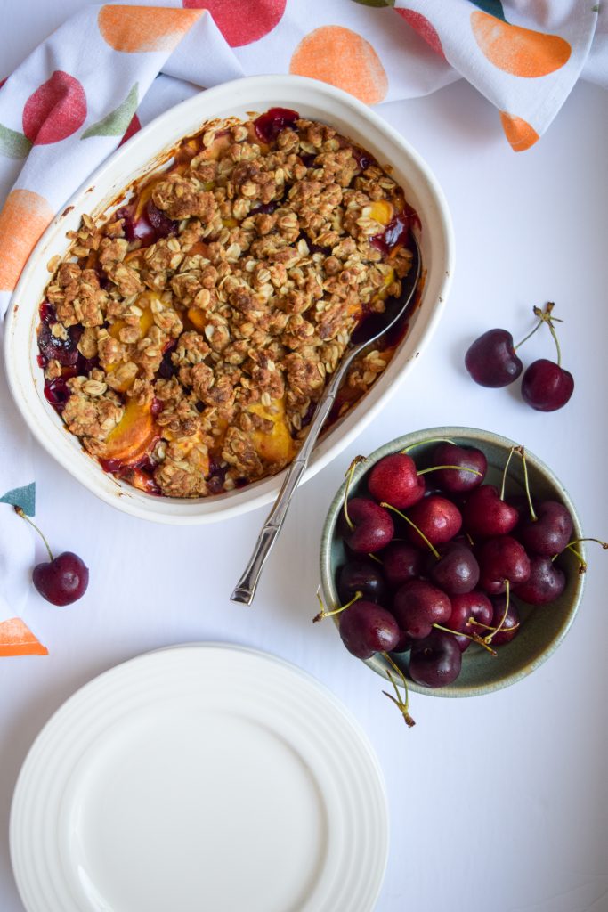 Peach cherry crisp in a white casserole dish on a white backdrop, surrounded by loose cherries, a green bowl full of cherries, white plates and a peach printed tea towel