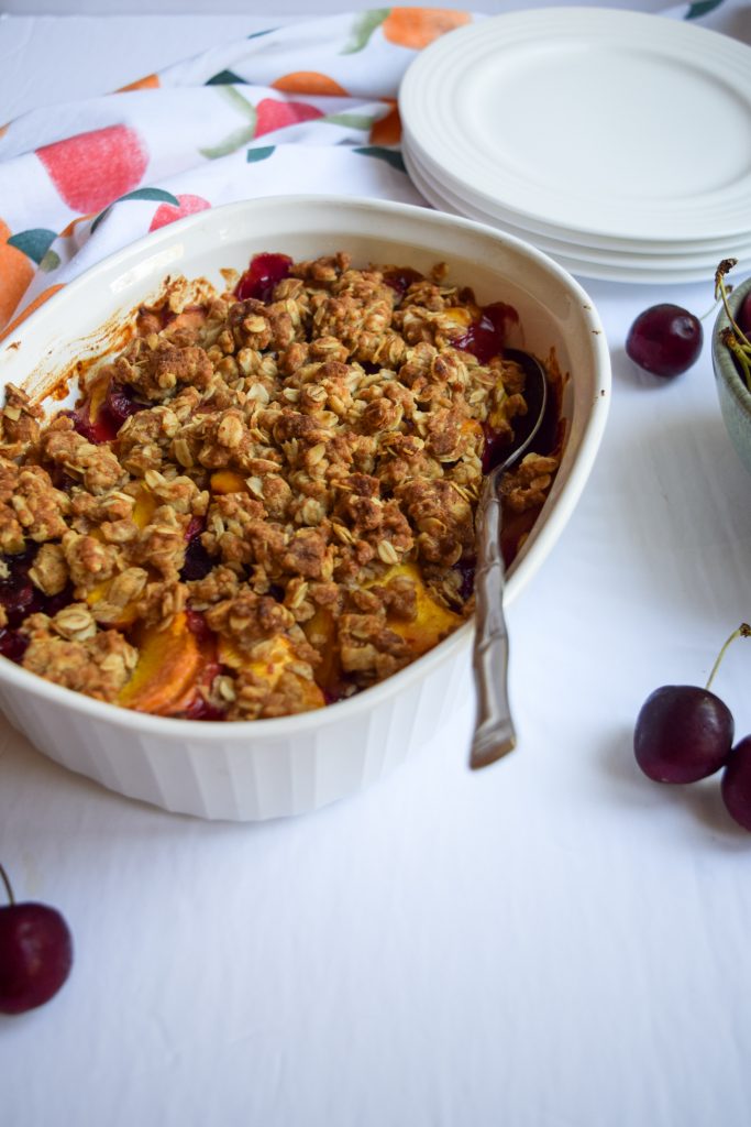 45 degree angle Peach cherry crisp in a white casserole dish on a white backdrop, surrounded by loose cherries, a green bowl full of cherries, white plates and a peach printed tea towel