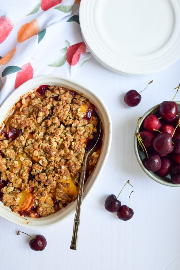 Peach cherry crisp in a white casserole dish on a white backdrop, surrounded by loose cherries, a green bowl full of cherries, white plates and a peach printed tea towel