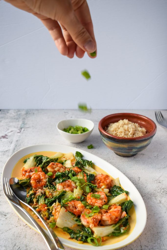 In-motion shot of a hand sprinkling green onions over an oblong plate of shrimp and bok choy. Behind a bowl of rice and a small bowl of green onions.