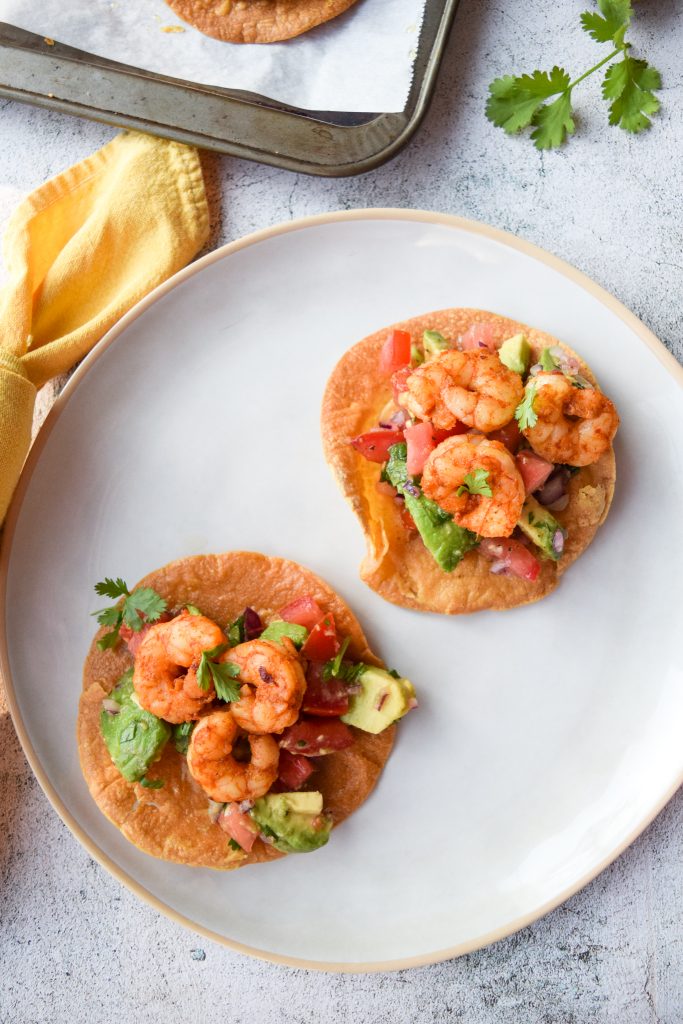 2 shrimp tostadas with avocado salad on a white plate, a yellow napkin to the left