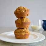 3 muffins stacked on top of each other on a white plate, glass of milk and blue glass in background