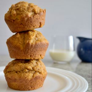 3 muffins stacked on top of each other on a white plate, glass of milk and blue glass in background