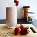 smoothie on a wooden cutting board, 2 strawberries and hemp seeds in the background, 2 jars in the background