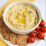 white bowl of hummus surrounded by crackers and cherry tomatoes, yellow napkin to left