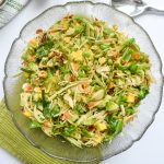 Green slaw in a glass bowl, atop a green napkin. Surrounded by white bowls, silver salad tongs