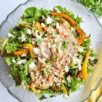 Glass salad bowl filled with salad and grilled veggies, topped with chicken and feta.