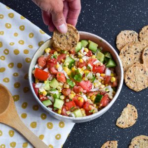 bowl of corn salsa with a hand and cracker dipping inside.