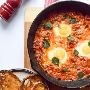 3 poached eggs in a tomato sauce, topped with parmesan cheese shavings and basil leaves in a cast-iron skillet