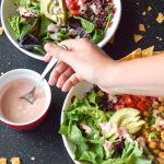 2 vegetarian taco bowls on a black backdrop, surrounded by broken tortilla chips. Arm reaches across to the left to spoon yogurt dressing.