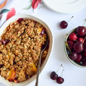 Peach cherry crisp in a white casserole dish on a white backdrop, surrounded by loose cherries, a green bowl full of cherries, white plates and a peach printed tea towel