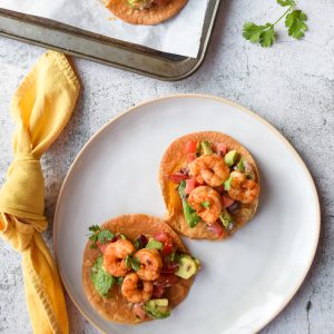 2 shrimp tostadas on a white plate, to the left a yellow napkin, above a parchment lined tray with another tostada