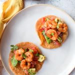 2 shrimp tostadas with avocado salad on a white plate, a yellow napkin to the left